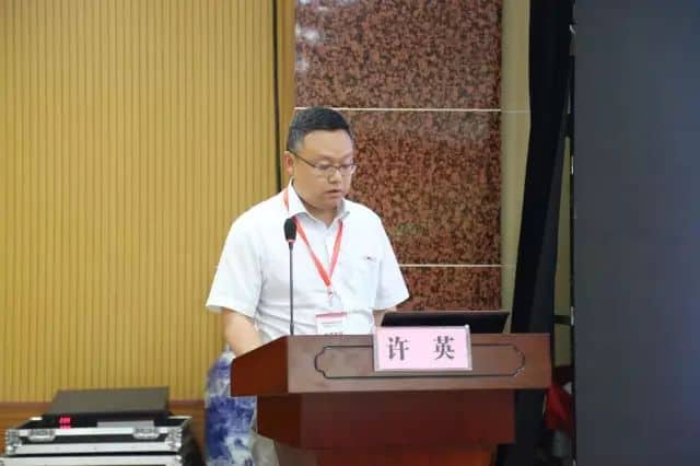 One participant wearing glasses stands behind a wooden lectern with a microphone and presentation screen, with a water bottle partly visible behind the lectern.