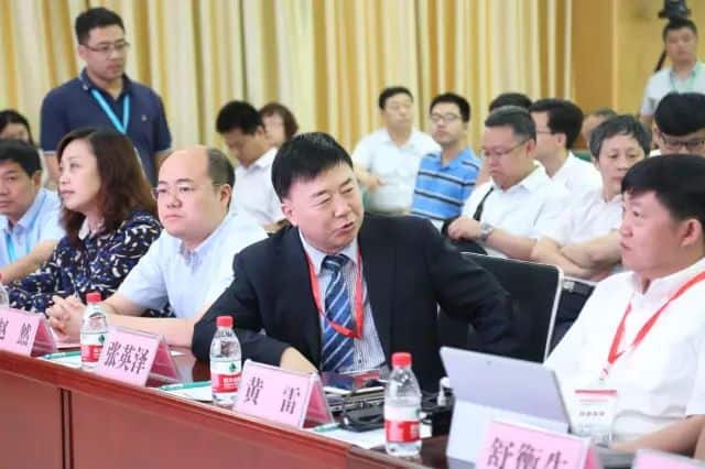 Approximately twenty adults sit in rows around conference tables with name placards, bottled drinks, laptops and papers, while one participant stands behind them.
