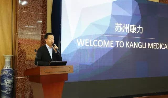 One speaker in a dark suit stands at a lectern with a microphone while a blue and purple presentation screen carries the English words KANGLI MEDICA and WELCOME TO KANGLI MEDICA.
