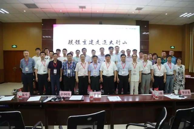 Roughly 28 adults stand in two rows behind a conference table with a projection screen, papers and name cards.