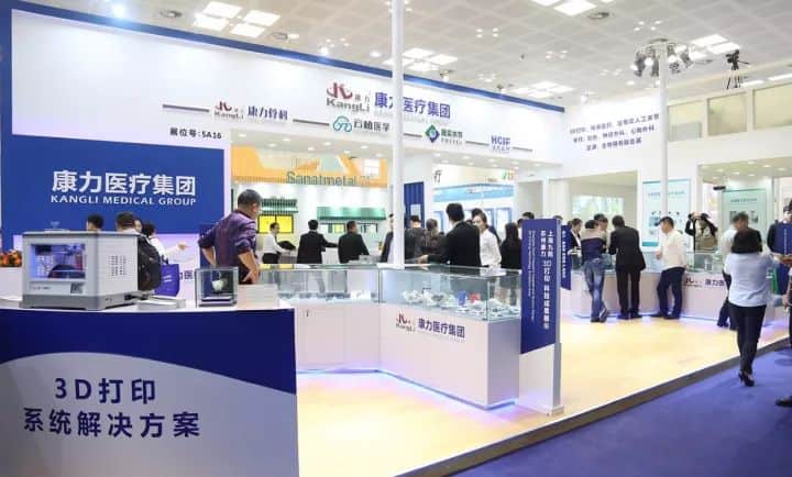About 20 visitors gather around a dark-blue and white exhibition stand with glass counters in a convention hall.