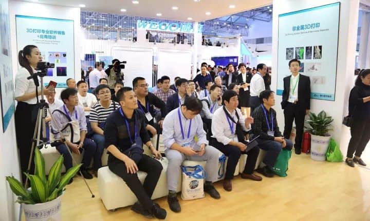 Approximately 30 people sit and stand around an exhibition-stand presentation, with a video camera on a tripod at left.