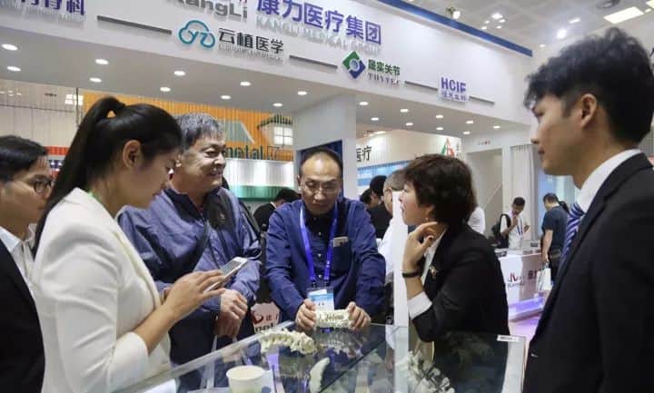Seven prominent visitors and additional attendees gather around a glass display counter at a convention stand.