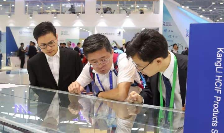 Three attendees lean over a glass display case in a busy exhibition hall while other visitors fill the background.