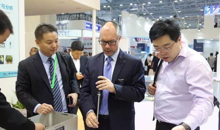 Three attendees stand at a foreground display in a busy trade-exhibition hall, with one holding an elongated object.