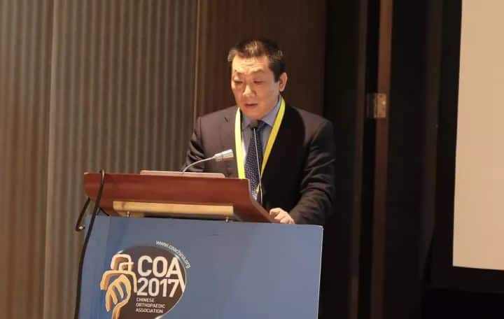 A presenter in a dark suit and yellow lanyard speaks behind a wooden lectern beside a projection screen.