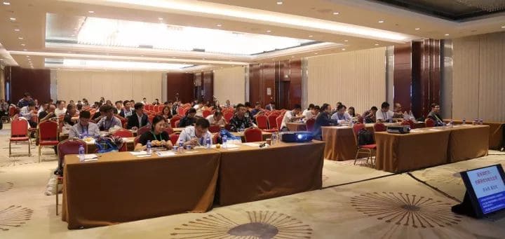 Approximately 80 people sit at long tables in a conference-room audience facing a presentation area.