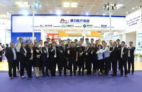 Approximately 30 participants in two rows pose with raised hands or closed fists in front of a branded exhibition stand.