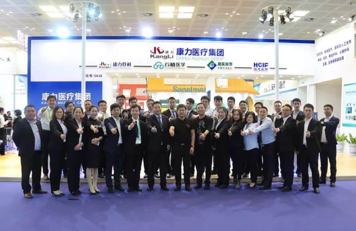 Approximately 30 participants in two rows pose with raised hands or closed fists in front of a branded exhibition stand.