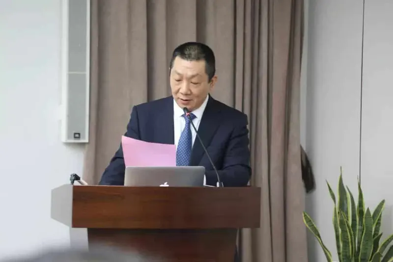 A speaker in a navy suit stands at a wooden lectern with a silver laptop and gooseneck microphone.