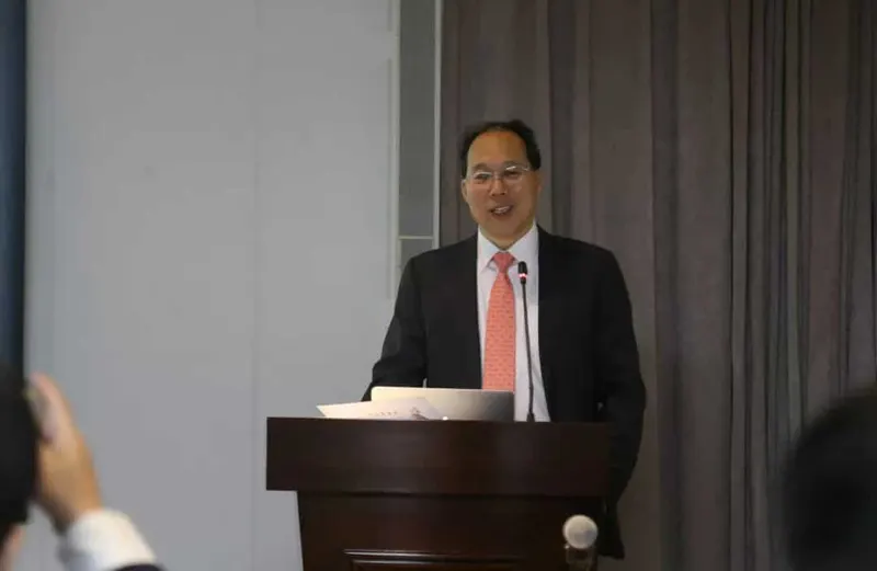 A speaker in a black suit and coral-pink patterned tie stands behind a wooden podium with a laptop.