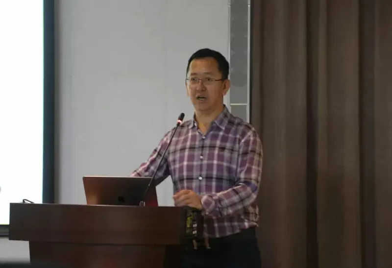 A speaker in a checked shirt stands at a wooden lectern beside a projection screen and microphone.