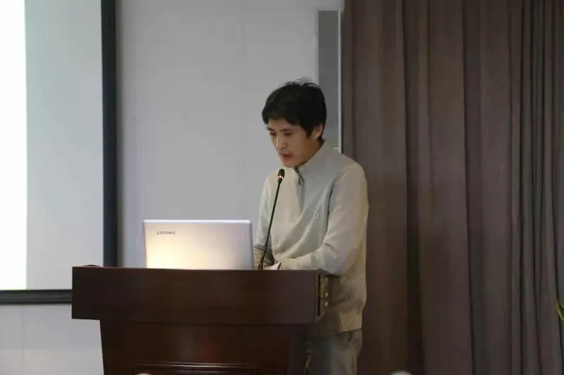 A speaker in a light grey sweater stands at a wooden lectern with a laptop and microphone while attendees appear at the bottom of the frame.