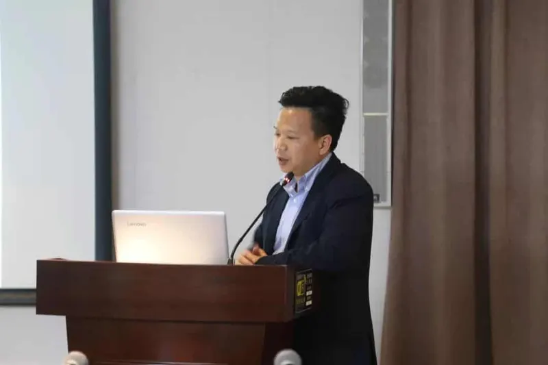A speaker in a dark jacket addresses the room from a wooden lectern with a laptop and microphone.