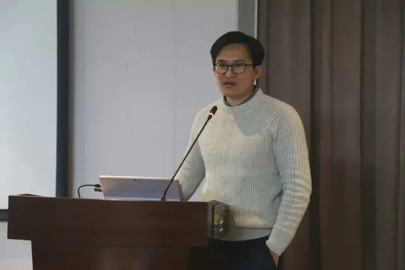 A speaker in a light grey sweater stands behind a wooden lectern with a laptop and microphone.