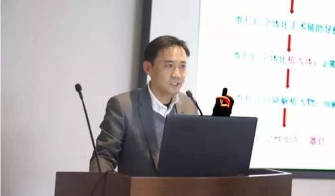 A speaker in a dark grey jacket stands behind a lectern with a laptop, two microphones, and a projected slide.