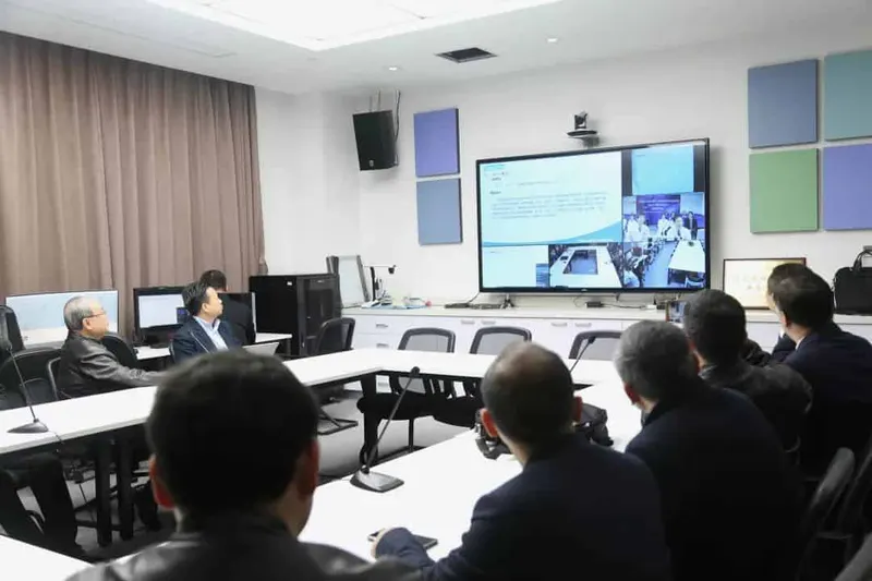 Six participants sit around a meeting room facing a wall display showing a presentation and video feeds.