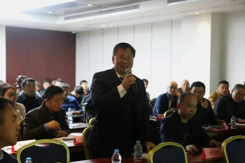 At least 20 participants sit at tables while a speaker in a dark suit addresses the room with a handheld microphone.