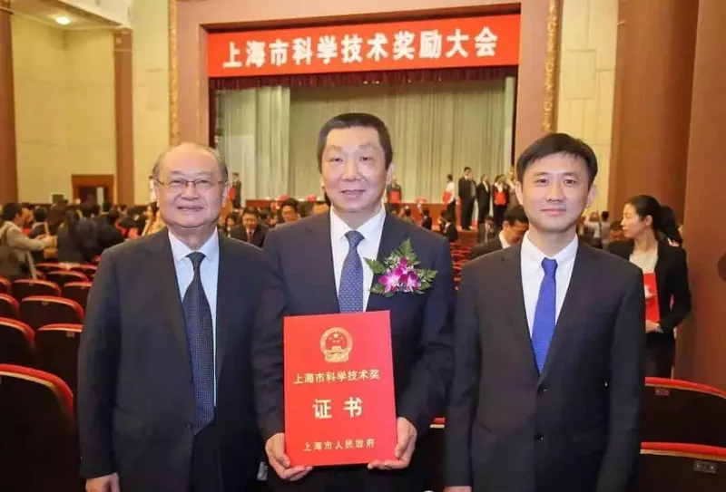 Three adults in dark suits stand in an auditorium, with one holding a red certificate and an audience behind them.