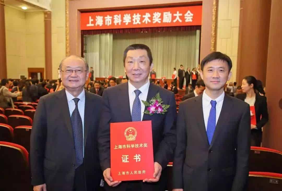 Three adults in dark suits stand in an auditorium, with one holding a red certificate and an audience behind them.