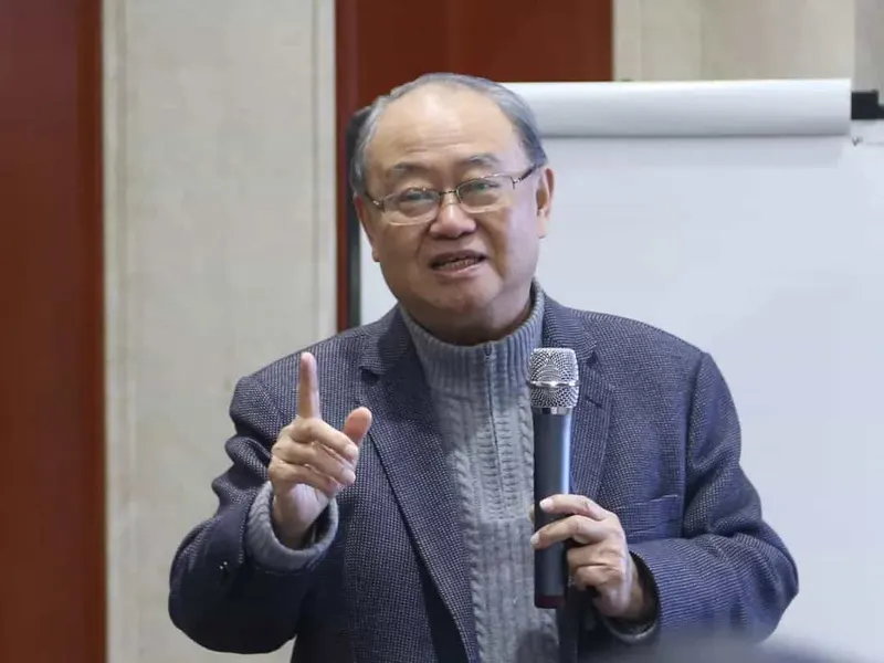 One speaker holds a microphone and raises an index finger beside a blank flip chart.