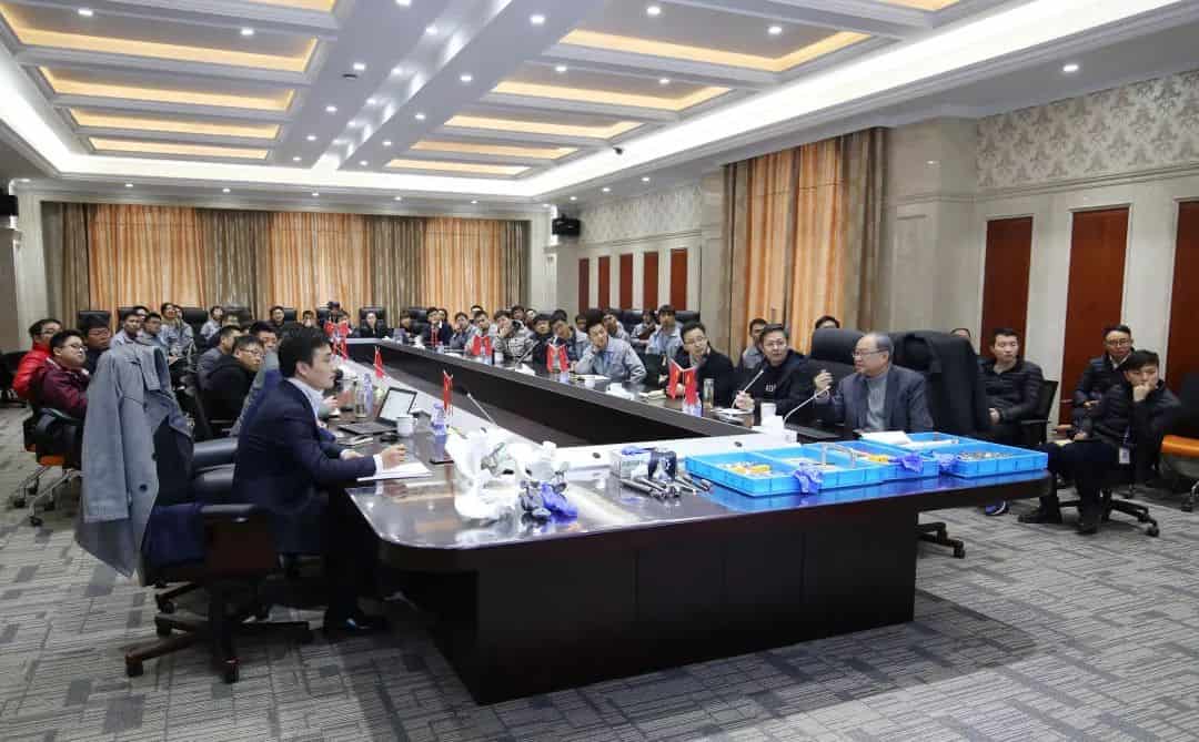 Approximately 35 participants sit around a long conference-room table with notebooks, microphones, and presentation materials.