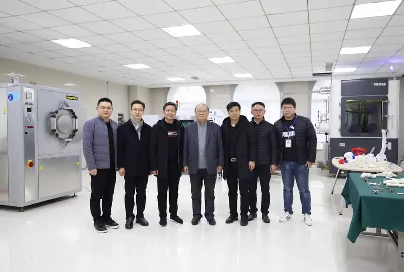 Seven people stand in a clean laboratory or production room beside industrial processing equipment and a worktable.