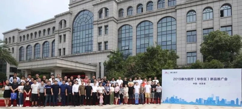 Approximately 45 adults and a child stand in rows outside a building beside a white event sign, with several participants holding bags.