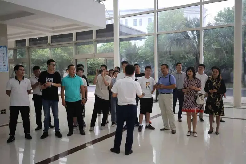 About 18 participants stand in a bright building lobby with large windows, while one person in the foreground faces the group.