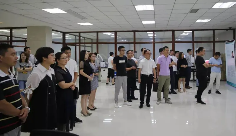 Approximately 25 to 30 people stand in a bright institutional room facing toward the right, with a tall white display panel nearby.