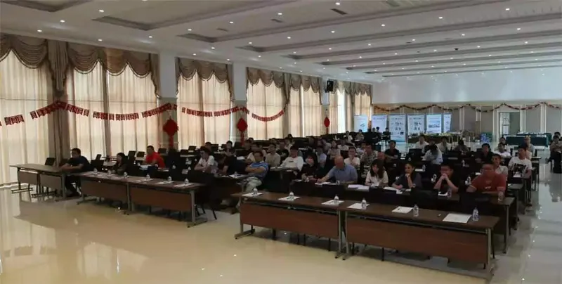 Approximately 70 to 90 attendees sit at rows of tables in a conference hall, with some using open laptops and others holding papers.