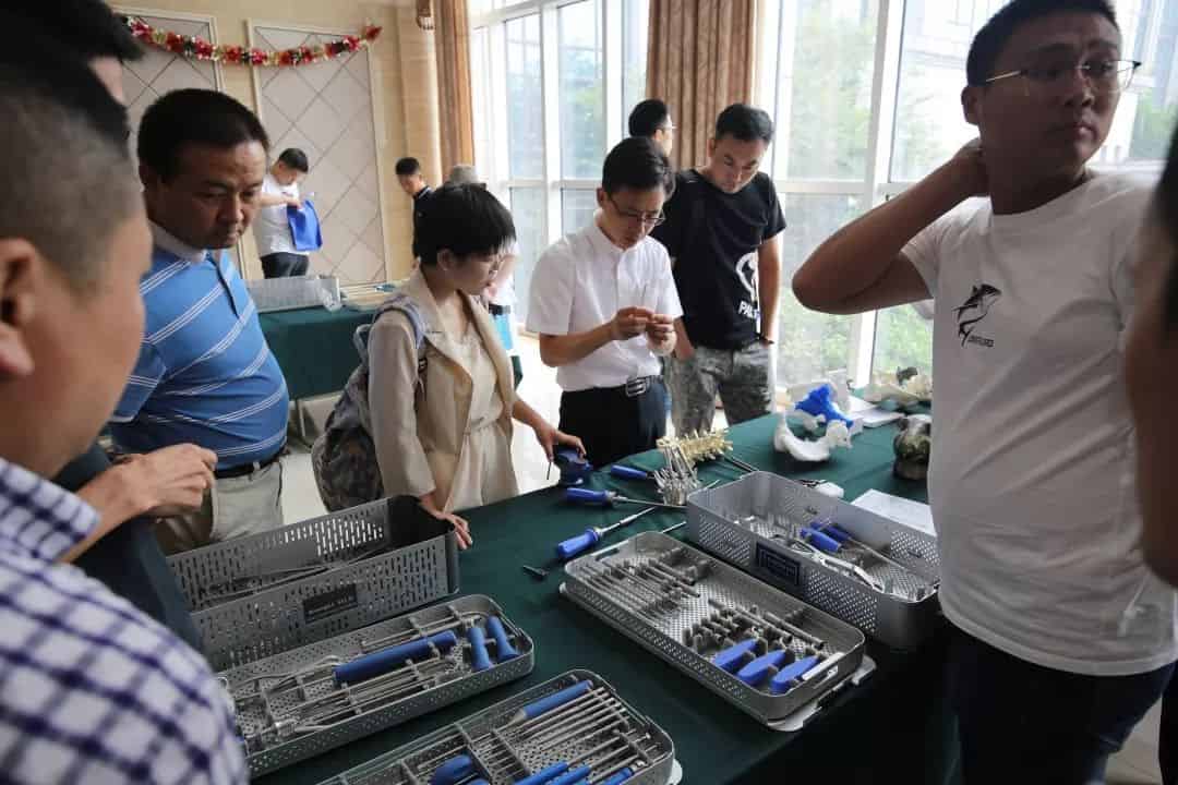 Nine people stand around tables covered with instrument trays and loose metal components in an indoor hall.
