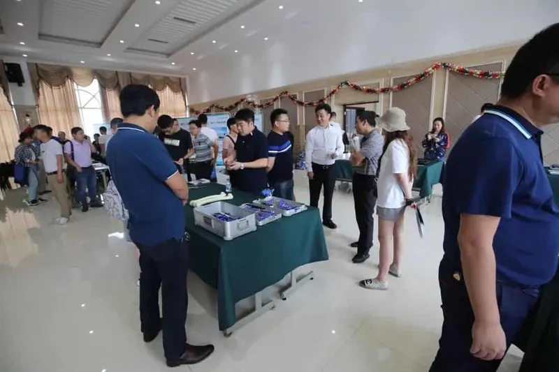 About 20 people gather around a central display table and trays of medical components in a crowded indoor hall.