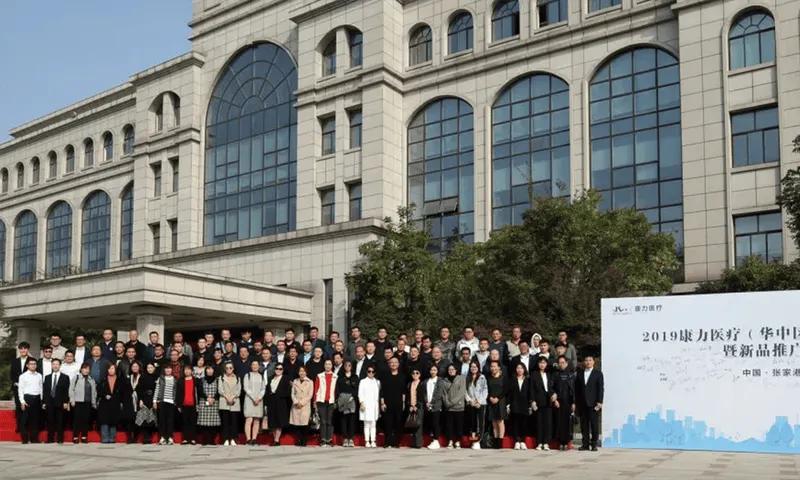 Approximately 65 people stand in several rows on a red carpet outside a formal building.
