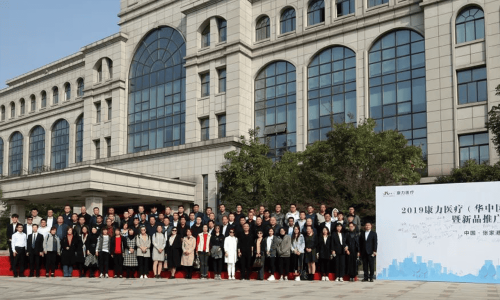 Approximately 65 people stand in several rows on a red carpet outside a formal building.