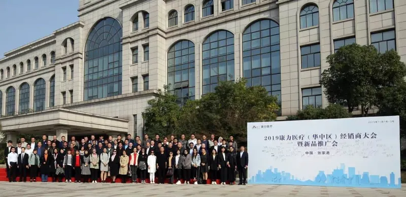 Approximately 70 people stand in several rows outdoors in front of a building and an event display board.