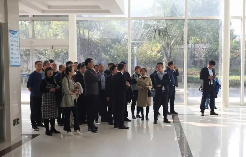 Approximately two dozen adults gather in a bright indoor lobby with glass walls and landscaping visible outside.