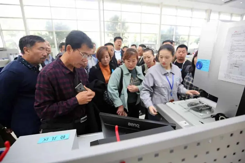 Approximately 18 to 20 adults gather around a large machine, computer monitor and technical sheet in an indoor factory area.