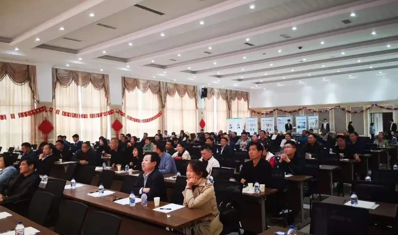 Approximately 70 people sit at long tables in rows in a conference room facing an unseen presentation area.