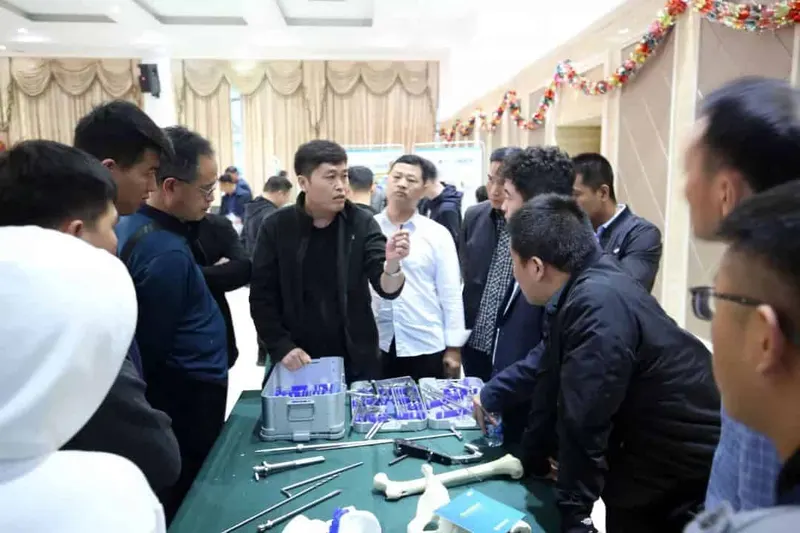 Approximately 14 people gather around a dark green-covered table displaying medical instruments and implant components in an indoor meeting room.