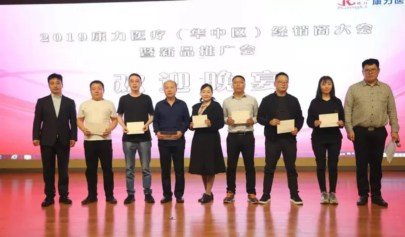 Nine people stand in a line on a conference stage in front of a projected presentation backdrop, with several holding papers or rectangular items.