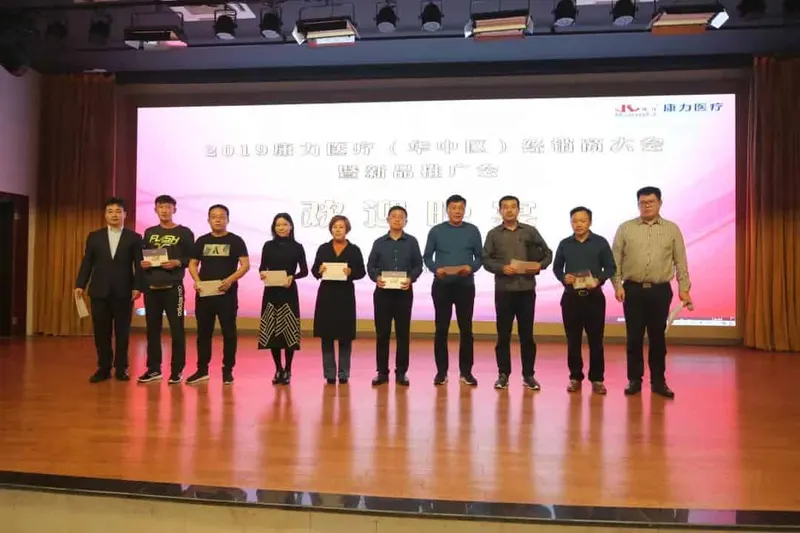 Ten people stand in a line on an auditorium stage in front of a projected event slide, with several holding documents, plaques or boxed items.