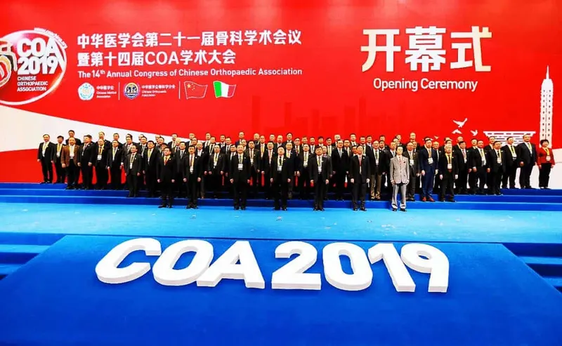 Approximately 60 participants in formal business attire stand in rows on a stage beneath a red conference backdrop.