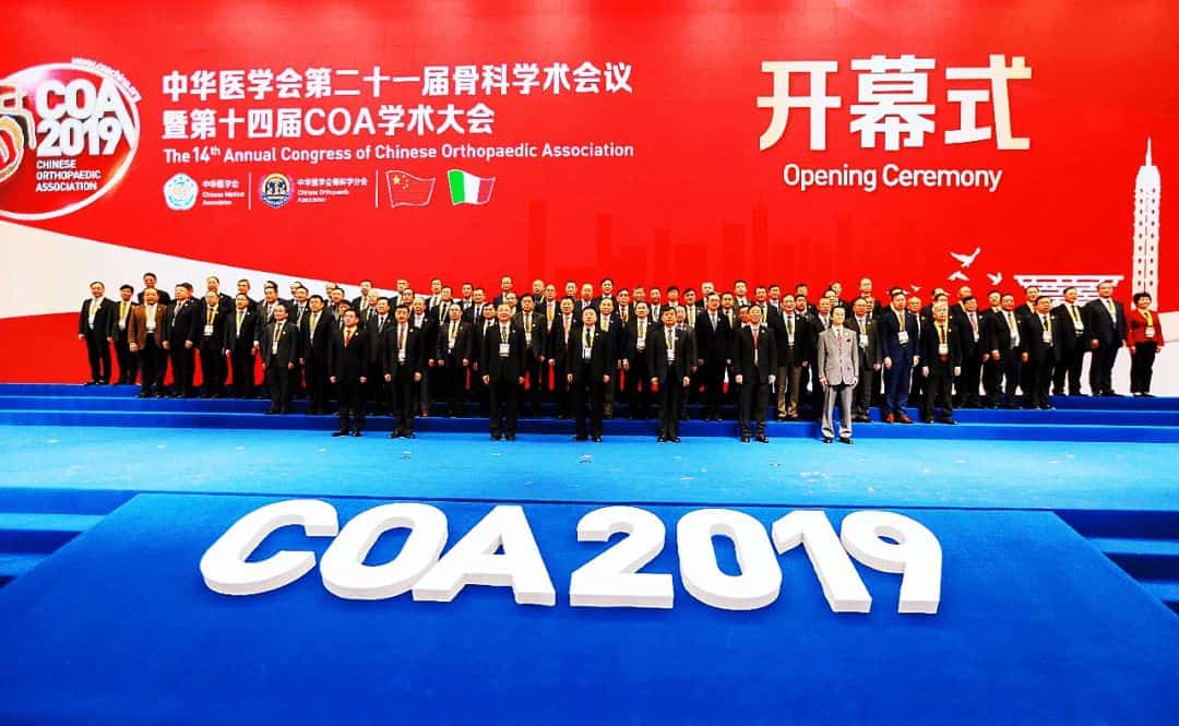 Approximately 60 participants in formal business attire stand in rows on a stage beneath a red conference backdrop.