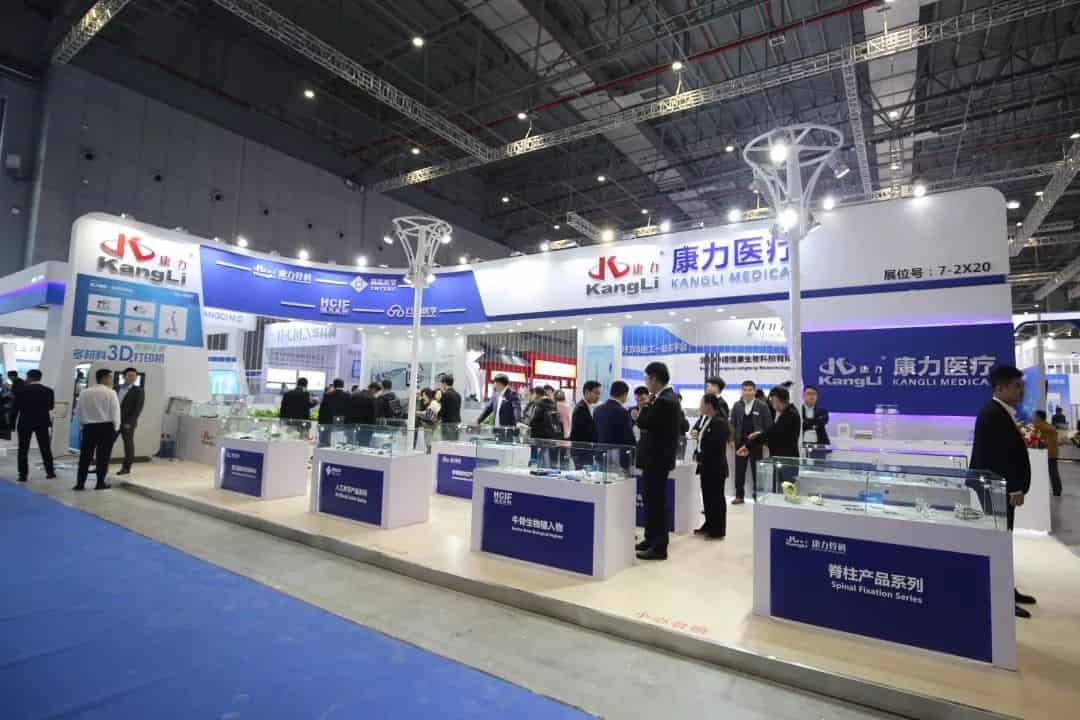 About 20 participants gather at a white-and-blue branded exhibition stand in a large convention hall.
