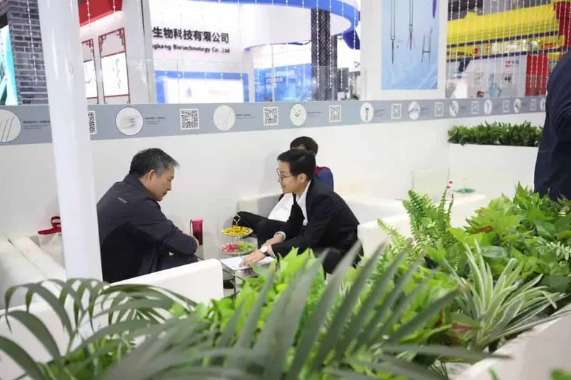 Four participants are visible at an exhibition booth, with two seated in white lounge chairs beside a low table.