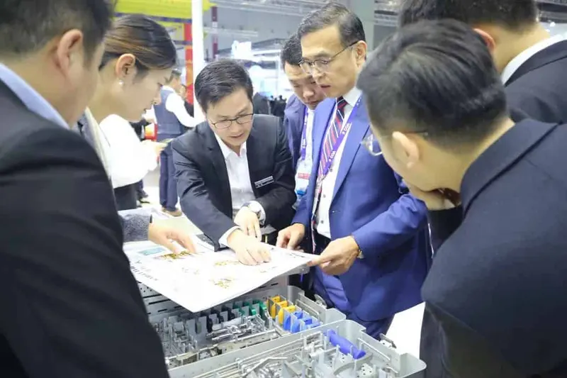 At least seven participants gather around an open gray compartmented case and a printed presentation sheet at an exhibition stand.