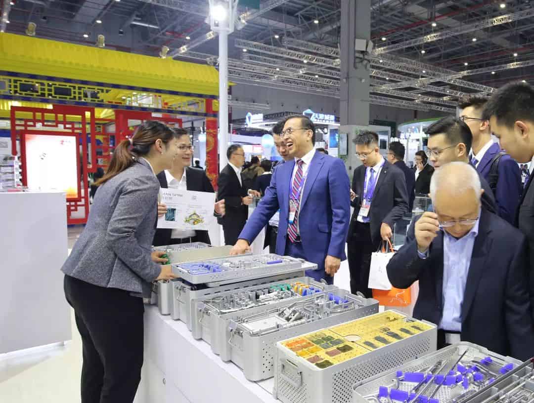 Approximately 12 participants gather around an exhibition counter in a convention hall, including a person in a bright blue suit.