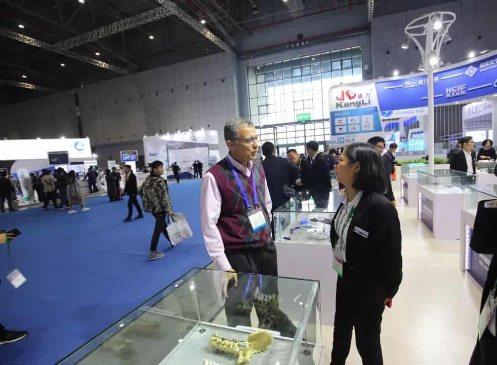 Two participants converse beside a display case in a blue-carpeted exhibition hall while visitors move between surrounding booths.