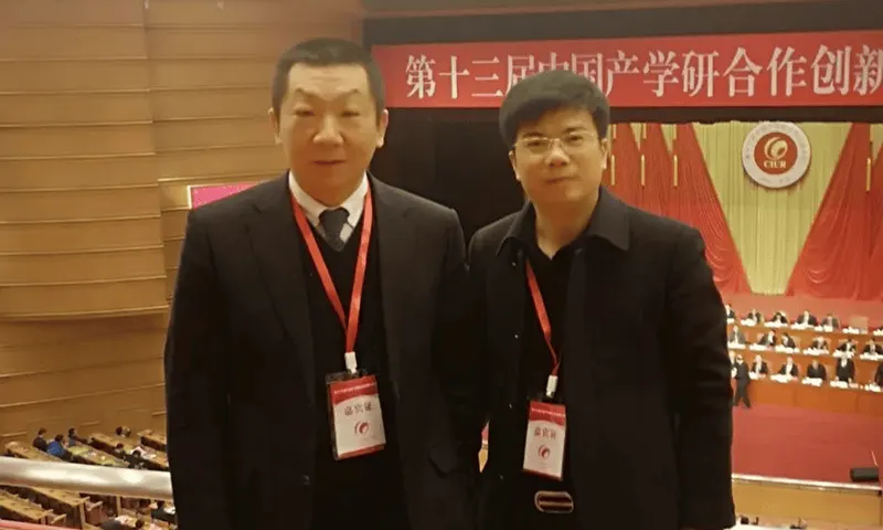 Two adults wearing red lanyards stand before a decorated stage in a conference hall with rows of seating behind them.